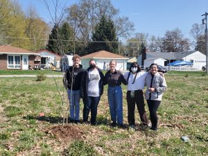 A group of young people stand in a field near a newly planted tree.