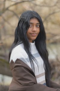 A portrait of a young woman with long black hair wearing informal neutral-color clothes, with brown trees in the background. 