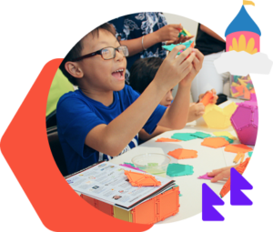 A young boy smiles holding a colored puzzle piece and sits at a table with colored pieces of a puzzle. 