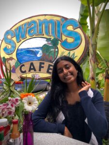 A picture of Sana Yooseph smiling, waving, seated at an outdoor cafe. 