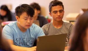 Two teen boys sit in front of a laptop computer.