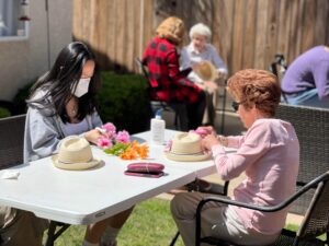 One female student, wearing a gray hoodie, is helping an elderly woman, wearing a pink shirt, to decorate a hat during the Springtime. It is outdoors and they are sitting at a table.