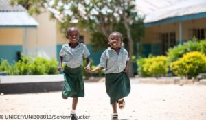wo girls in green skirts and green and white checked shirts run through a school courtyard.