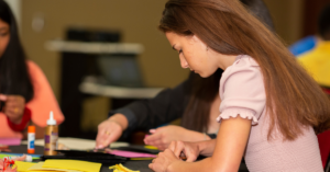A side profile photo of a young woman working with craft items on a table