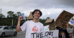 a young person stands, smiling, holding a bullhorn in the air and a cardboard sign with a drawing of planet Earth.