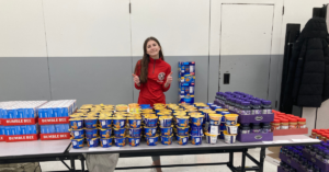 A young woman in a red school athletic shirt stands in front of a table with stacks of boxed foods.