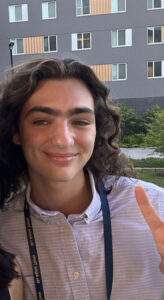 A young person smiles at the camera in front of a grey building