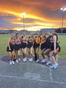 Aspen poses in a line hugging several other cheerleaders in front of an empty sports field and a brilliant orange and yellow sunset.