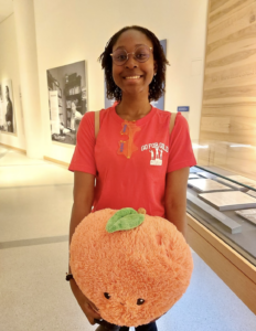 Chikanma grins while holding a large plush peach and wearing a bright T-shirt that says "go for gold"