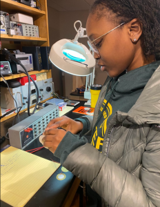 Chikanma looking through a large magnifying glass light at a desk with piezoelectric equipment 