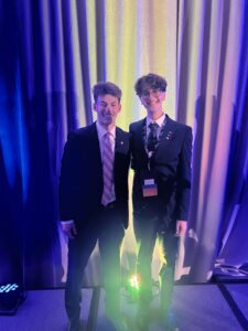 Moe and another student stand together wearing suits and ties in front of a stage curtain and lights.