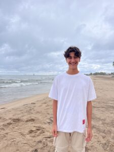 Moe stands smiling on a beach on a cloudy day.