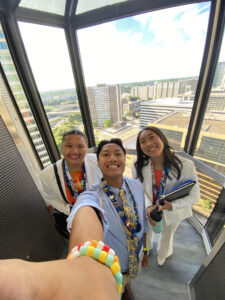 Three young people pose for a selfie in an elevator. Nicole stands with her two siblings who are dressed professionally and smiling while wearing lanyards