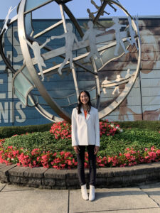 Nicole, wearing a white blazer and boots, stands in front of the Kiwanis International office building’s metal globe sculpture.