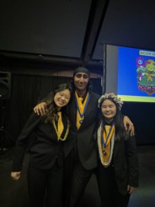 Sunny stands with his arm around two other students wearing bright gold ties, yellow and blue braided ribbon leis and black suits.  
