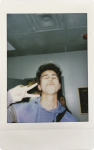 A polaroid vintage-style photo of Armin holding up a peace sign and making a funny face with his eyes closed  