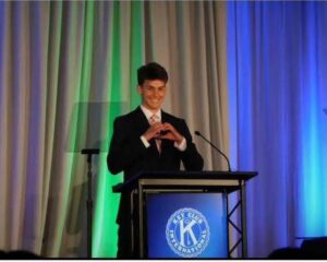 David smiles, holding his hands together to form a heart, while standing behind a podium with the Key Club seal. 