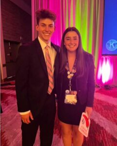 David and another student stand smiling together, wearing suits in front of a brightly lit stage curtain.