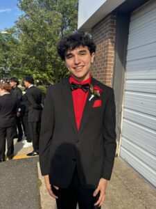 Kevin stands, smiling, wearing a black suit and bowtie with a red shirt and rose boutonniere 