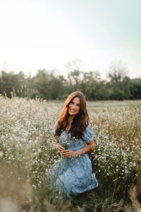 Carissa kneels in a field of little white wildflowers wearing a light blue, summery dress 