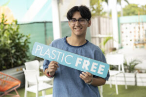 A student smiles holding a sign that reads in big block letters plastic free.