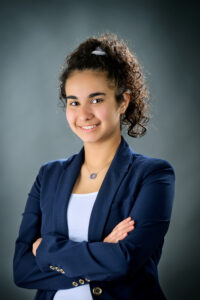 A professional headshot photo of someone with a friendly smile and a curly ponytail wearing a navy blazer.