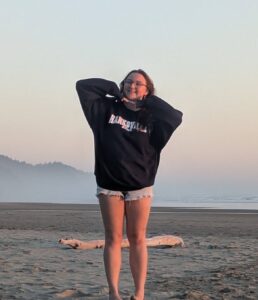 3. A young person grins wearing cutoff shorts, a oversized black sweatshirt on an empty beach in front of a large piece of driftwood