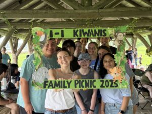 A group of young people smile posing behind a green handmade frame that reads I-I Kiwanis family picnic 2025