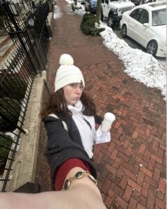 A selfie photo of a young person holding a paper cup, wearing a white sock cap and scarf standing on a brick sidewalk lined with snow.
