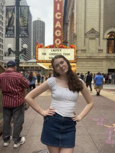 A young person stands in a city, hands on hips, wearing a denim skirt and a white tank top in front of a marquee that reads Laufey The Goddess tour.