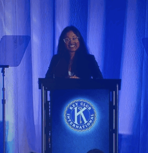 a young person stands smiling wearing a black blazer behind a podium with a Key Club seal on it