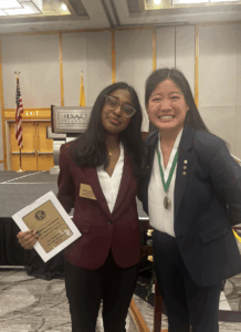 Two young people stand arm in arm wearing business outfits. One is holding a framed award and the other is wearing a gold medal around her neck