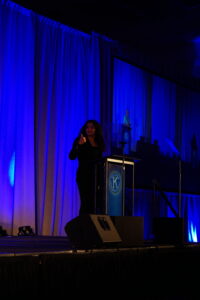 On a big stage, someone stands behind a podium with a Key Club seal and gives the thumbs-up sign.