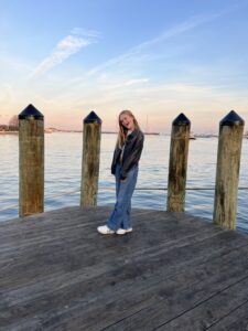 A young person grins on a dock with peaceful water and a nice skyline behind her.