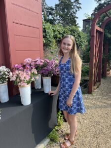 A young person smiles standing outside next to several white containers of pink and purple flowers.