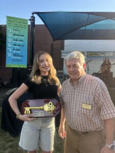 Two people pose for a photo, one is a young person holding an award with a large golden key on a wooden plaque. The other is an older person wearing a name tag and smiling. 