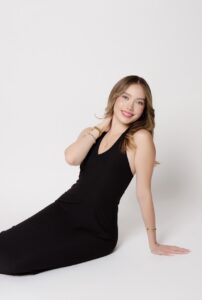A young person smiles calmly sitting elegantly in front of an all white background wearing a black dress 