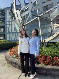 Two young people stand outdoors arm in arm in front of a metal structure wearing business outfits 