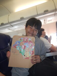 A young person grins holding a large card while siting in an airplane