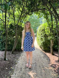 A young person stands under a canopy of trees wearing a blue summer dress.