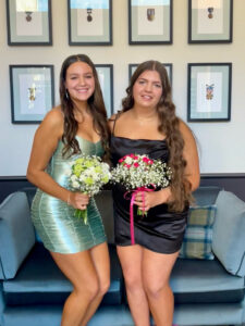 Two young people stand arm in arm wearing cocktail dresses holding flowers