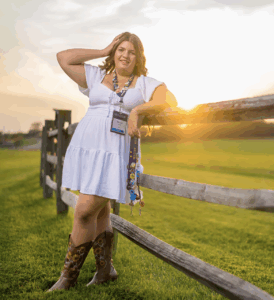 A young person wearing a white summer dress and cowboy boots leans on a rail fence in the country with a stunning sunset behind