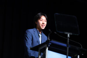 a young person stands in a spotlight behind a podium getting ready to speak