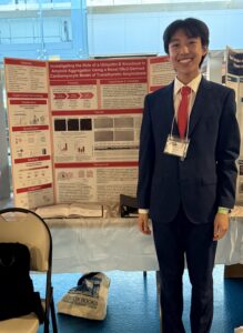 A young person wearing a black suit with a red tie stands in front of a trifold display 