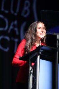 A young person stands behind a podium wearing a red blazer