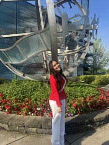 A young person wearing business clothing stands in front of a flowerbed and a metal global structure