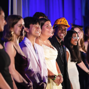 A line of young people beaming smiles wearing formal clothing with one wearing a yellow construction hat with signatures in black marker