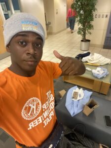 A selfie photo of a young person holding a thumbs up wearing an orange Key Club t-shirt and grey Key Club stocking cap