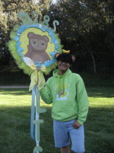 A young person wearing a bright green hoodie a fun headband stands holding a sign round sign of a teddy bear bordered with ribbon, tulle and the letters D28S