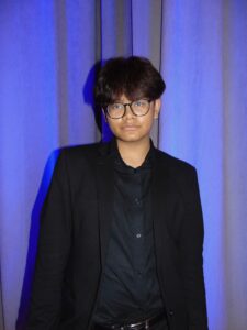 A young person strikes a serious pose wearing a black suit in front of lit curtains
